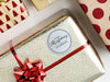 A white glitter cardstock gift tag with black ink is shown on a Christmas present. The present is wrapped in gold paper and has a red bow and ribbon around it. The background of the picture is a white surface.