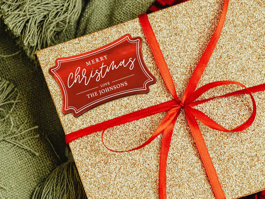 A red foil cardstock gift tag is shown on a glitter wrapped box. The box also has a red ribbon on it. The box is seen on a green blanket surface.