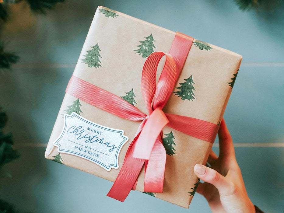 A white glitter cardstock gift tag with blue ink is shown on a Christmas present. The present is wrapped in Christmas tree paper and has a red ribbon around it. The background of the picture is a green/blue.