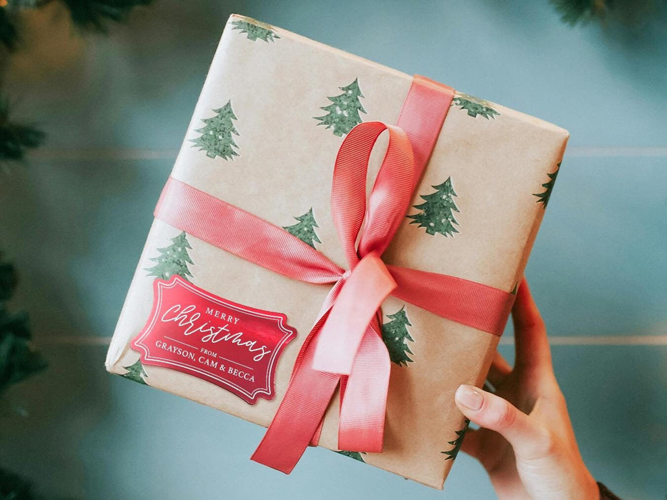 A red foil cardstock gift tag is shown on a Christmas present. The present is wrapped in Christmas tree paper and has a red ribbon around it. The background of the picture is a green/blue.