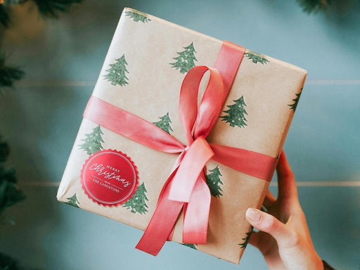 A red foil cardstock gift tag is shown on a Christmas present. The present is wrapped in Christmas tree paper and has a red ribbon around it. The background of the picture is a green/blue.