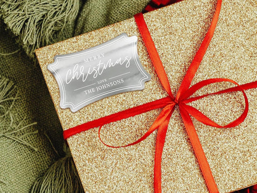 A silver foil cardstock gift tag is shown on a glitter wrapped box. The box also has a red ribbon on it. The box is seen on a green blanket surface.