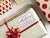 A white glitter cardstock gift tag with red ink is shown on a Christmas present. The present is wrapped in gold paper and has a red bow and ribbon around it. The background of the picture is a white surface.