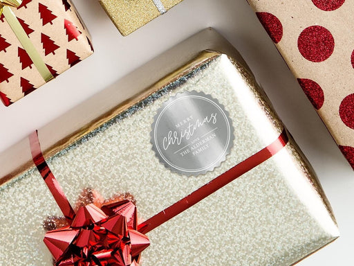 A silver foil cardstock gift tag is shown on a Christmas present. The present is wrapped in gold paper and has a red bow and ribbon around it. The background of the picture is a white surface.