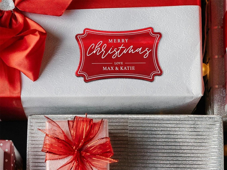 A red foil cardstock gift tag is shown on a Christmas present. The present is wrapped in white paper and has a red bow and ribbon around it. It has other Christmas presents surrounding it.