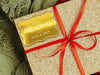 A gold foil cardstock gift tag is shown on a glitter wrapped box. The box also has a red ribbon on it. The box is seen on a green blanket surface.