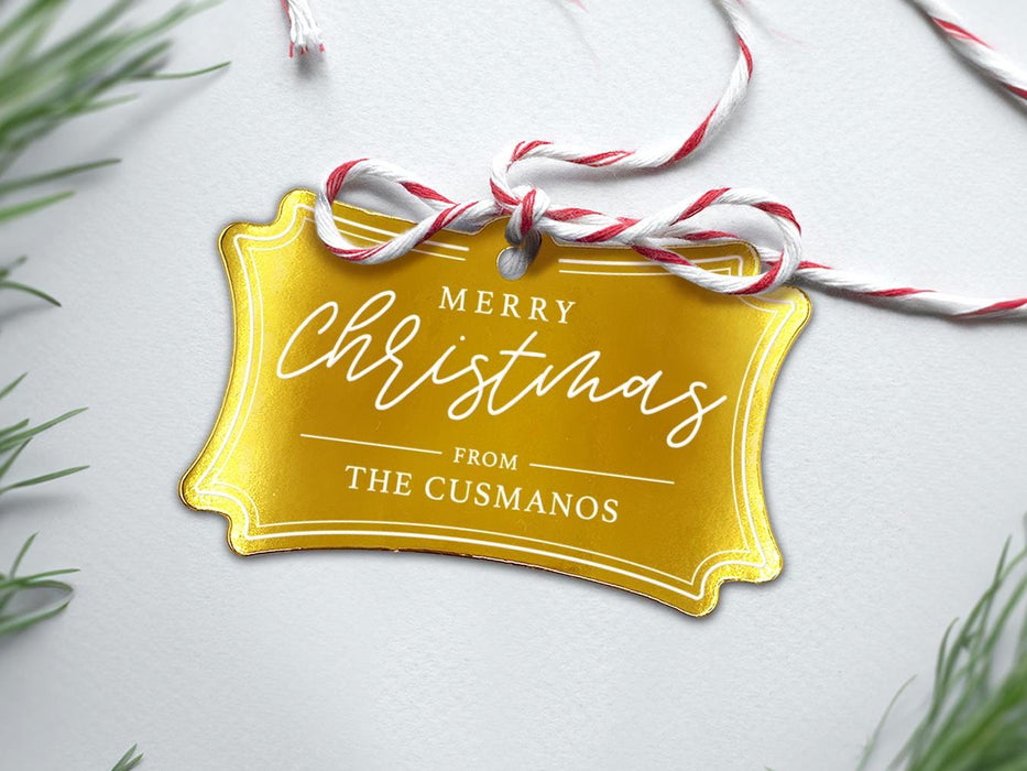 A gold foil cardstock Merry Christmas gift tag is shown sitting on a white background surrounded by Christmas tree clippings. The tag has a red and white string looped through the top of it.