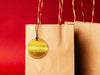 A gold foil cardstock Merry Christmas gift tag is shown hanging from a brown, craft paper gift bag. The bag is on a red backdrop.