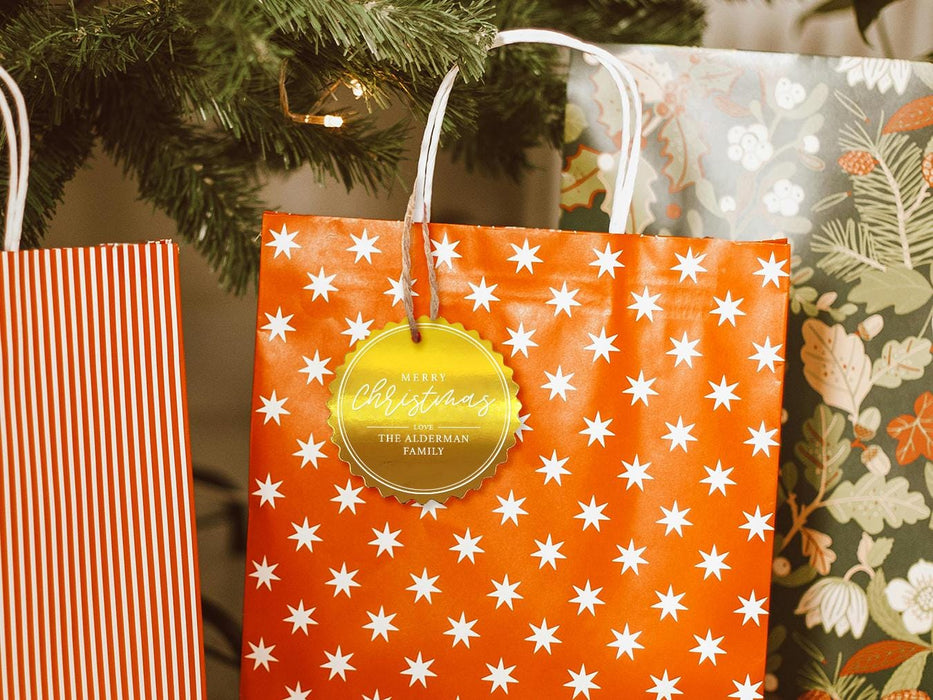 A gold foil cardstock Merry Christmas gift tag is shown hanging from a star-patterned white and red gift bag. The bag sits under a Christmas tree surrounded by other presents.