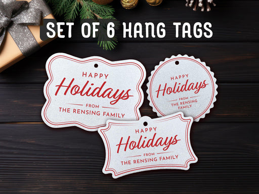 Text reads, Set of 6. Three white glitter cardstock Happy Holidays gift tags printed in red ink are shown on a dark wooden surface. Pine tree branches and a Christmas present are shown around the tags.