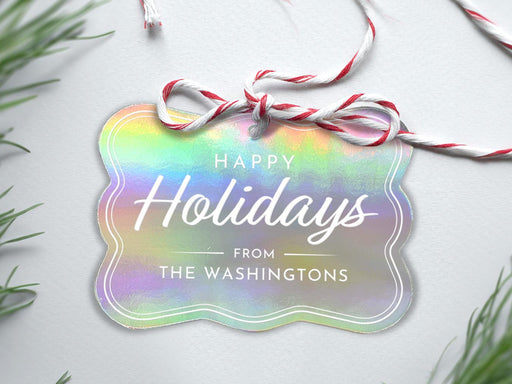 An iridescent foil cardstock Happy Holidays gift tag is shown sitting on a white background surrounded by Christmas tree clippings. The tag has a red and white string looped through the top of it.