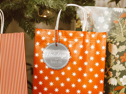 A silver foil cardstock Happy Holidays gift tag is shown hanging from a star-patterned white and red gift bag. The bag sits under a Christmas tree surrounded by other presents.