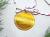 A gold foil cardstock Merry Christmas gift tag is shown sitting on a white background surrounded by Christmas tree clippings. The tag has a red and white string looped through the top of it.