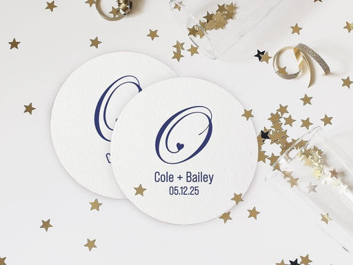 Two coasters lay on white surface with champagne glasses and gold stars surrounding them. Coasters feature a heart initial design with a monogram, couples names, and wedding date.