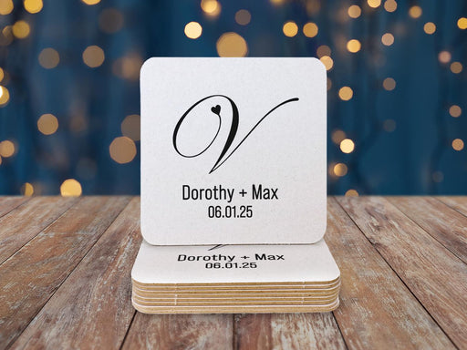 A stack of coasters sit on a wooden surface. Coasters feature a heart initial design with a monogram, couples name, and wedding date.