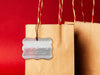 A silver foil cardstock Merry Christmas gift tag is shown hanging from a brown, craft paper gift bag. The bag is on a red backdrop.