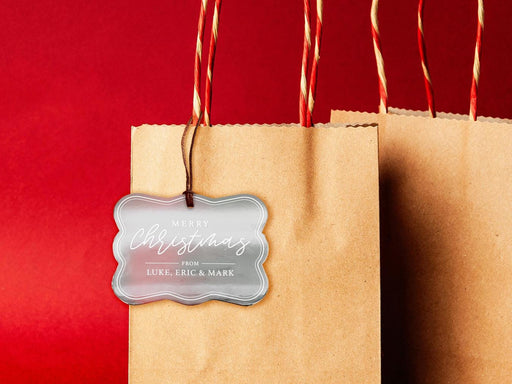 A silver foil cardstock Merry Christmas gift tag is shown hanging from a brown, craft paper gift bag. The bag is on a red backdrop.