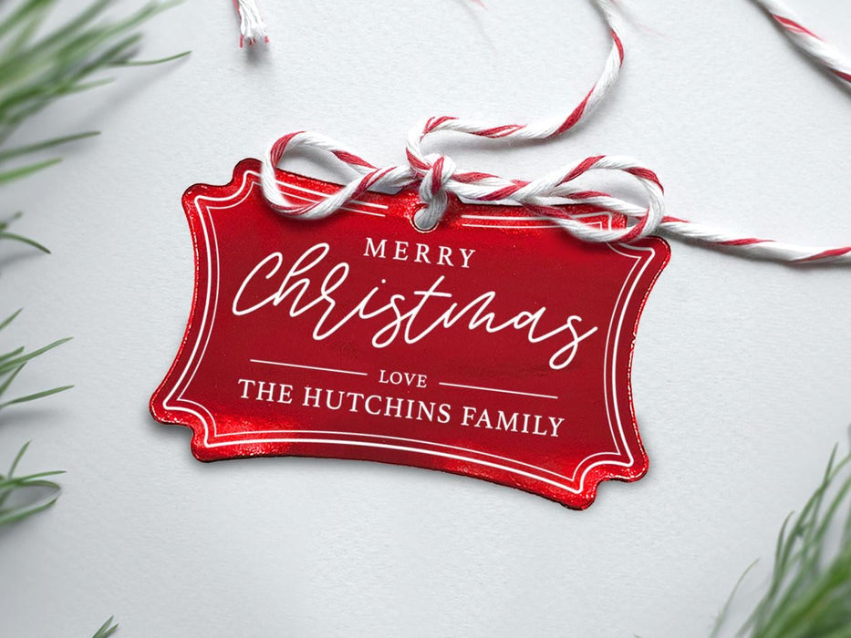 A red foil cardstock Merry Christmas gift tag is shown sitting on a white background surrounded by Christmas tree clippings. The tag has a red and white string looped through the top of it.