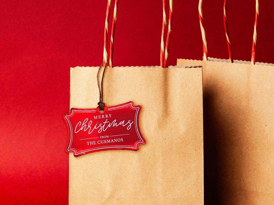 A red foil cardstock Merry Christmas gift tag is shown hanging from a brown, craft paper gift bag. The bag is on a red backdrop.