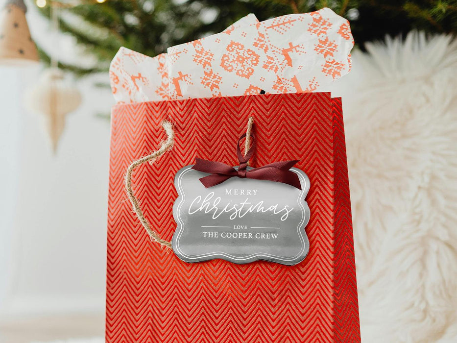 A silver foil cardstock Merry Christmas gift tag is shown hanging from red gift bag. The bag sits under a Christmas tree against a white background.