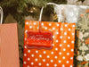 A red foil cardstock Merry Christmas gift tag is shown hanging from a star-patterned white and red gift bag. The bag sits under a Christmas tree surrounded by other presents.