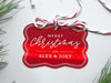 A red foil cardstock Merry Christmas gift tag is shown sitting on a white background surrounded by Christmas tree clippings. The tag has a red and white string looped through the top of it.