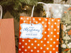 A white glitter cardstock Merry Christmas gift tag with blue ink is shown hanging from a star-patterned white and red gift bag. The bag sits under a Christmas tree surrounded by other presents.