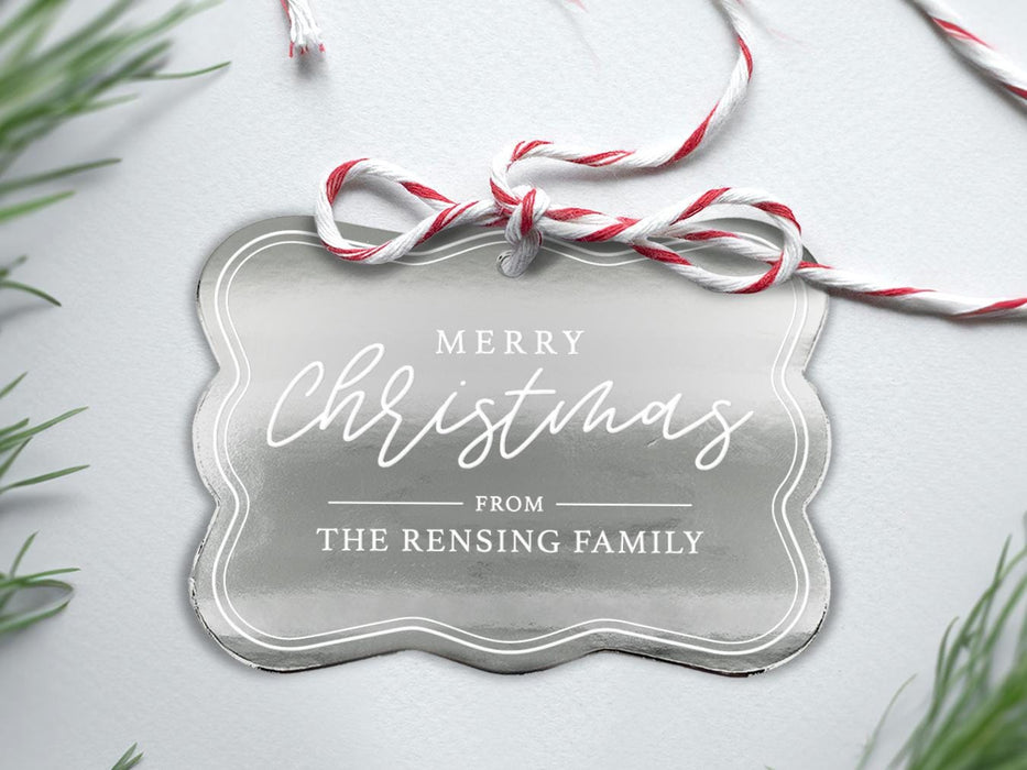 A silver foil cardstock Merry Christmas gift tag is shown sitting on a white background surrounded by Christmas tree clippings. The tag has a red and white string looped through the top of it.