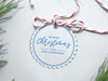 A white glitter cardstock Merry Christmas gift tag with blue ink is shown sitting on a white background surrounded by Christmas tree clippings. The tag has a red and white string looped through the top of it.