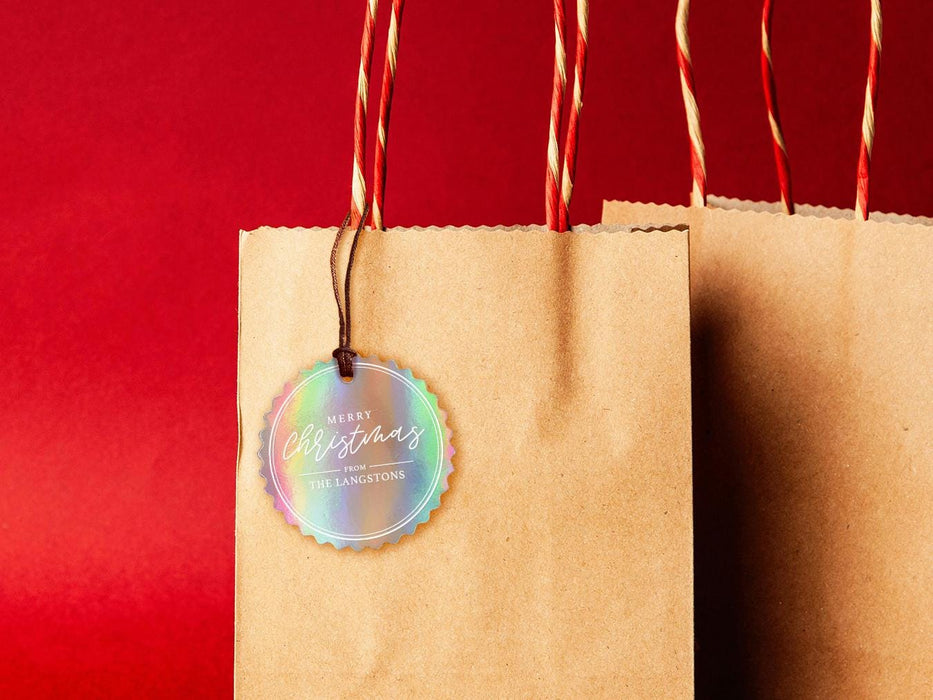 An iridescent cardstock Merry Christmas gift tag is shown hanging from a brown, craft paper gift bag. The bag is on a red backdrop.