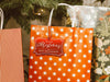 A red foil cardstock Merry Christmas gift tag is shown hanging from a star-patterned white and red gift bag. The bag sits under a Christmas tree surrounded by other presents.