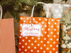 A white glitter cardstock Merry Christmas gift tag with red ink is shown hanging from a star-patterned white and red gift bag. The bag sits under a Christmas tree surrounded by other presents.