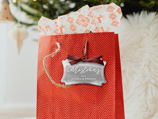 A silver foil cardstock Merry Christmas gift tag is shown hanging from red gift bag. The bag sits under a Christmas tree against a white background.