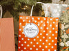 A white glitter cardstock Merry Christmas gift tag with black ink is shown hanging from a star-patterned white and red gift bag. The bag sits under a Christmas tree surrounded by other presents.