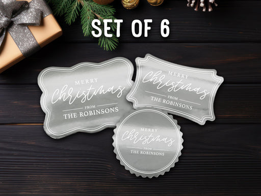 Text reads, Set of 6. Three silver foil cardstock Merry Christmas gift tags are shown on a dark wooden surface. Pine tree branches and a Christmas present are shown around the tags.