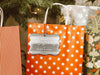 A silver foil cardstock Merry Christmas gift tag is shown hanging from a star-patterned white and red gift bag. The bag sits under a Christmas tree surrounded by other presents.