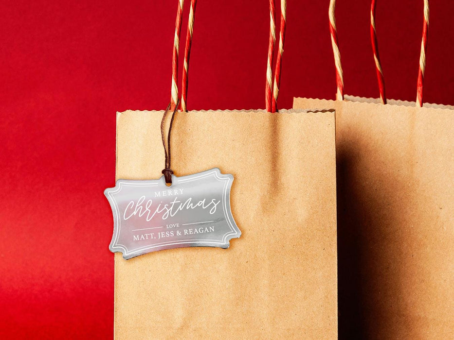 A silver foil cardstock Merry Christmas gift tag is shown hanging from a brown, craft paper gift bag. The bag is on a red backdrop.