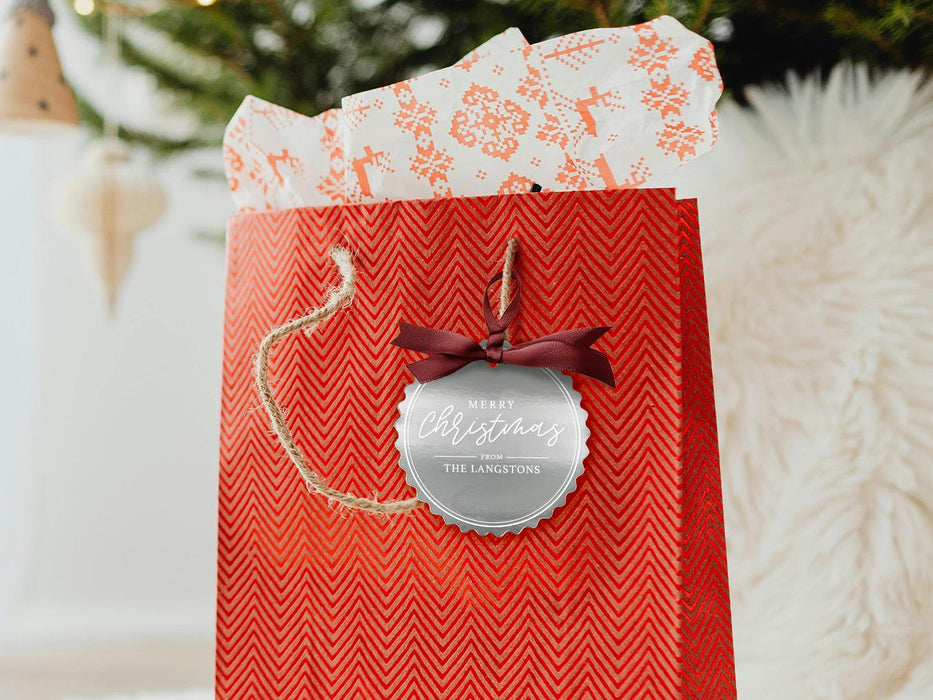 A silver foil cardstock Merry Christmas gift tag is shown hanging from red gift bag. The bag sits under a Christmas tree against a white background.