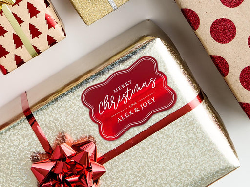 A red foil cardstock gift tag is shown on a glitter wrapped box. The box also has a red ribbon on it. The box is seen on a white background surrounded by other Christmas gifts.