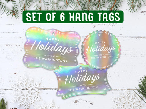 Text reads, Set of 6 Hang Tags. Six iridescent foil cardstock Happy Holidays hang tags are shown on a white wooden surface. Pine tree branches and snowflakes surround the tags.