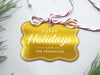 A gold foil cardstock Happy Holidays gift tag is shown sitting on a white background surrounded by Christmas tree clippings. The tag has a red and white string looped through the top of it.