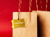 A gold foil cardstock Happy Holidays gift tag is shown hanging from a brown, craft paper gift bag. The bag is on a red backdrop.