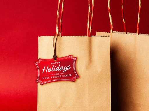 A red foil cardstock Happy Holidays gift tag is shown hanging from a brown, craft paper gift bag. The bag is on a red backdrop.