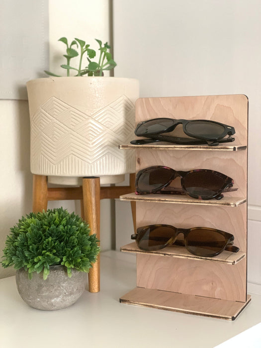 wooden glasses stand with multiple sunglasses sitting on a countertop surrounded by multiple potted plants