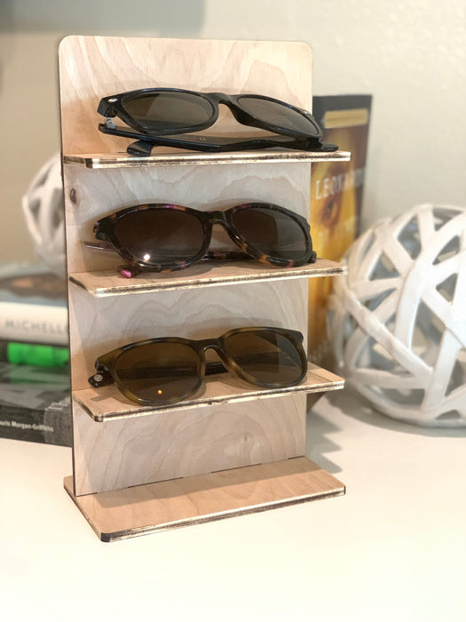 wooden glasses stand with multiple sunglasses sitting on a countertop surrounded by books and decor