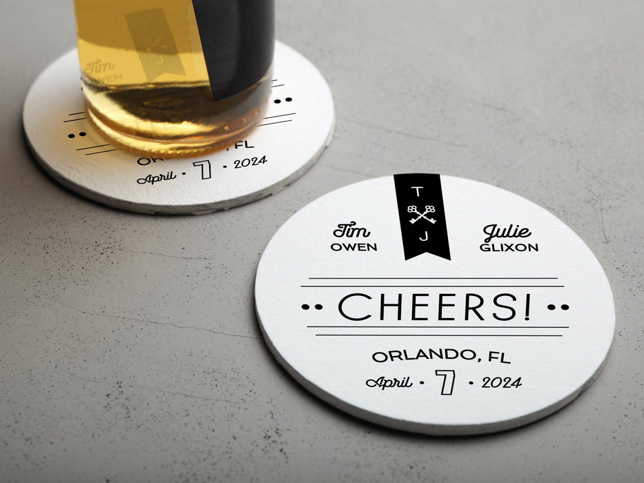 One coaster has a drink on it and an empty coaster sits beside it. Coasters feature CHEERS! Modern Wedding design. Coasters show married couple names and first name initials, the word “CHEERS!”, and the wedding location and date. Design is printed in black on a white coaster. These coasters also have decorative keys, lines, and dots on it.