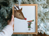 hand holding a wooden frame with a christmas print of a mouse decorating a tree in front of pine trees