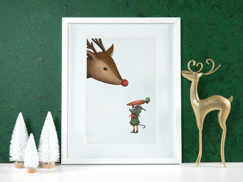 white frame with christmas print of an elf mouse offering a carrot to a reindeer against a green background ontop of a white counter surrounded by holiday decor such as white pine trees and a golden sparkly reindeer