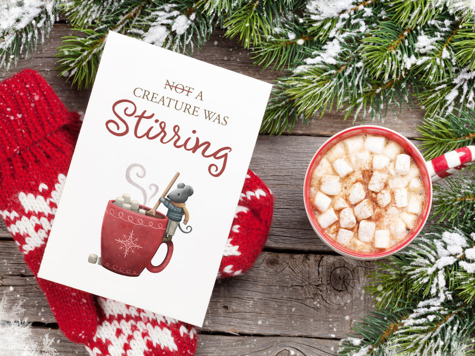 single print of mouse stirring artwork ontop of wooden table with red and white mittens, hot chocolate with marshmallows, and pine leaves covered with snow