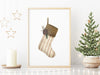 gold framed Christmas print with a rustic stocking surrounded by gold holiday decor such as a candle, gold stars, and a mini tree decorated with stars and mini lights